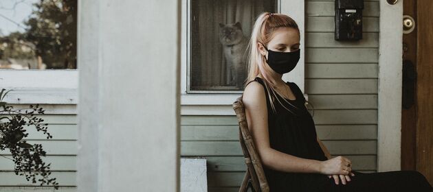 Eine junge Frau mit einer Mund-Nase-Bedeckung sitzt auf einer Terrasse und schaut traurig nach unten