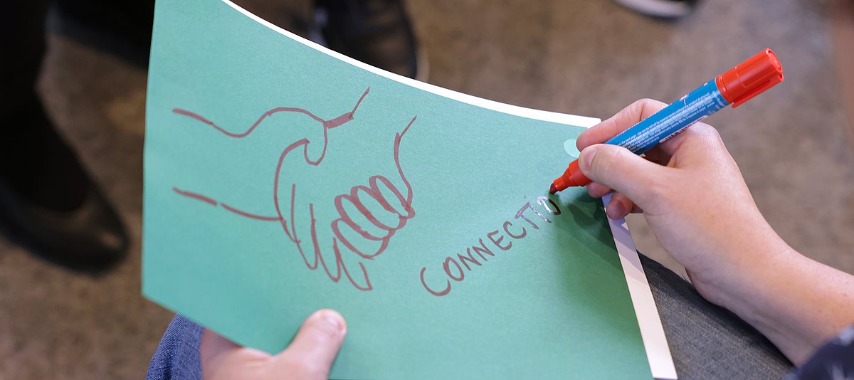 Eine Hand schreibt das Wort Connection auf ein Blatt, auf dem Blatt sind zwei gezeichnete Hände, die sich umfassen.