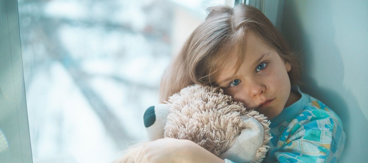 Ein Kind sitzt an einem Fenster und schaut traurig in die Kamera, im Arm hält es ein Kuscheltier