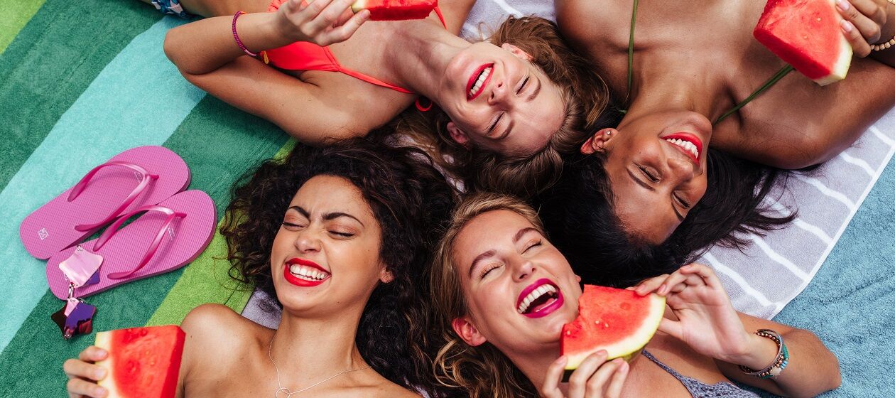Eine Gruppe junger Frauen in Badekleidung von oben fotografiert lacht mit Melonenschnitzen in der Hand