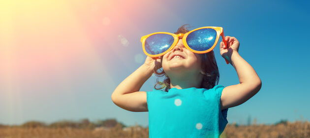Ein Kind mit übergroßer Sonnenbrille schaut in den sonnigen Himmel.