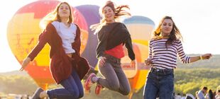 Drei junge Frauen springen in die Luft, hinter ihnen sind Heißluftballons zu sehen