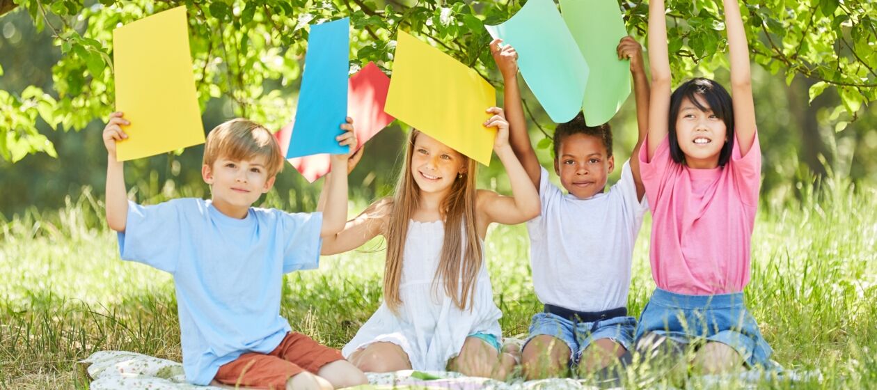 Vier Kinder sitzen draußen auf einer Wiese und halten verschiedenfarbige Zettel in die Luft