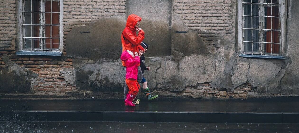 Eine Mutter in roter Regenjacke mit zwei Kindern an der Hand geht im Regen auf nassem Bürgersteig vor desolatem Haus.