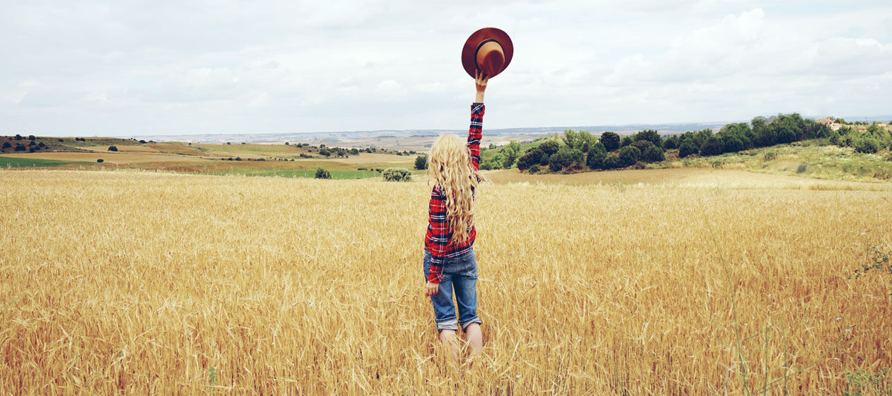 Ein Mädchen steht auf einem großen Weizenfeld und hält eine Hut hoch in die Luft