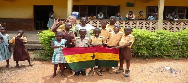 Eine Gruppe Kinder steht mit der Freiwilligen Marlene Bayer von weltwärts vor einer Schue. Sie halten die Flagge Ghanas vor sich.