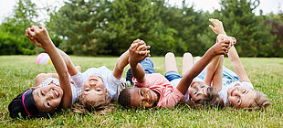 Ein Gruppe von Kindern unterschiedlicher Herkunft liegen auf einer Wiese und halten sich an den Händen und lächeln.