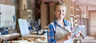Eine junge Frau steht lächelnd in einer Holzwerkstatt und hält ein tablet in der Hand.