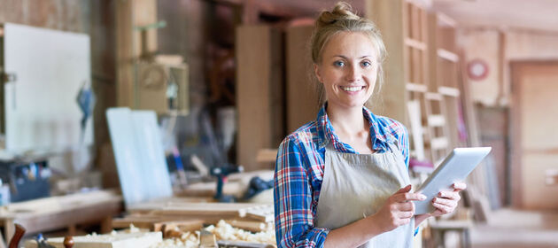 Eine junge Frau steht lächelnd in einer Holzwerkstatt und hält ein tablet in der Hand.