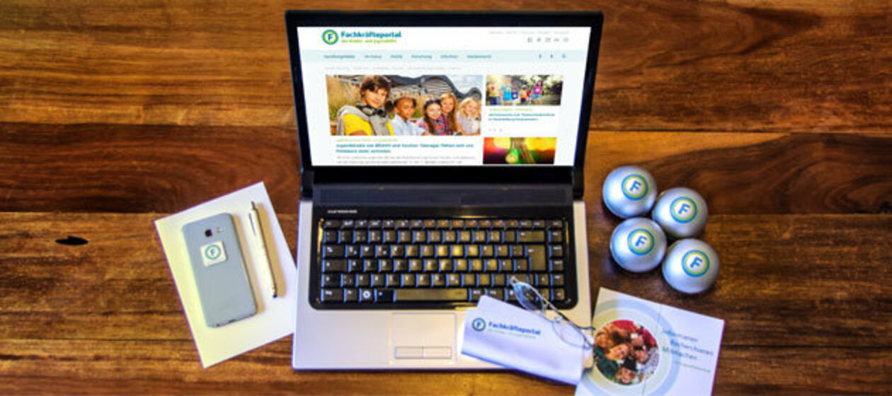Ein Laptop mit der Startseite des Jugendhilfeportals steht mit Werbemitteln auf einem braunen Holztisch. 