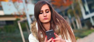 Eine Schülerin ist alleine draußen unterwegs und schaut traurig auf ihr Smartphone und schreibt etwas