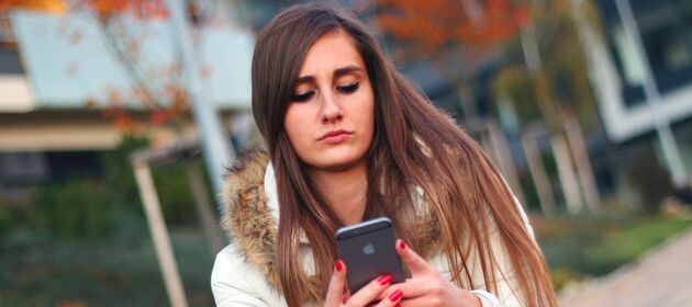 Eine Schülerin ist alleine draußen unterwegs und schaut traurig auf ihr Smartphone und schreibt etwas