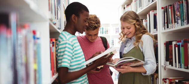 Drei Jugendliche stehen zwischen Bücherregalen und lesen in Büchern
