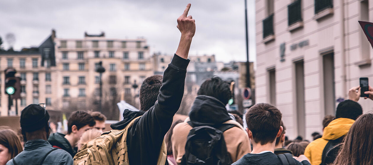 Junge Erwachsene demonstrieren in einer Stadt. In der Bildmitte streckt ein Demonstrant den Mittelfinger aus.