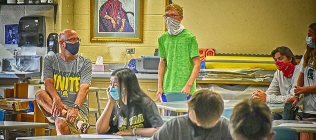 Schüler/-innen und ein Lehrer beim Unterricht während der Coronapandemie, mit Abstand und Mund-Nasen-Schutz