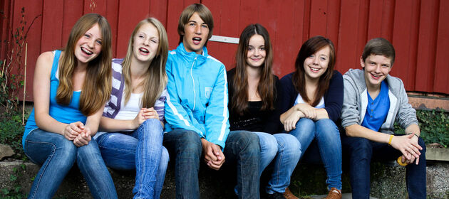 Eine Gruppe Jugendlicher sitzen vor einer roten Holzwand und lachen in die Kamera. 