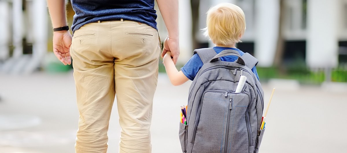 Ein Vater hält seinen kleinen Sohn an der Hand, der einen überdimensionalen Rucksack trägt