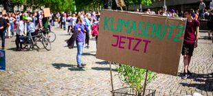 Auf einem Pappschild steht „Klimaschutz jetzt“. Im Hintergrund die Teilnehmenden einer Demonstration in sommerlicher Stadt.