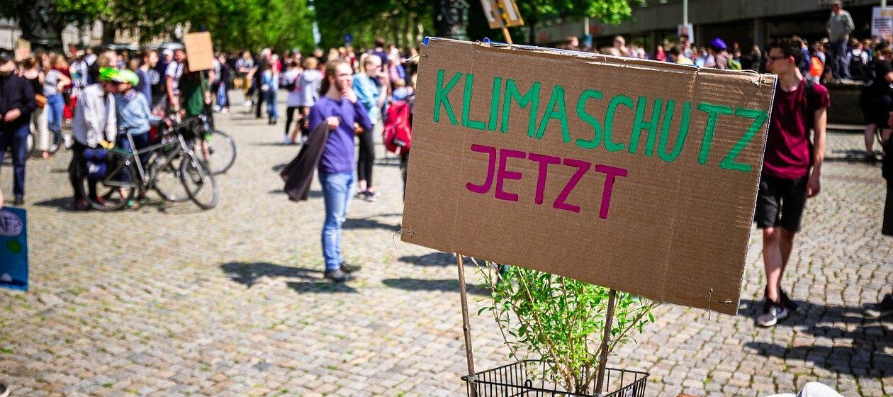 Auf einem Pappschild steht „Klimaschutz jetzt“. Im Hintergrund die Teilnehmenden einer Demonstration in sommerlicher Stadt.