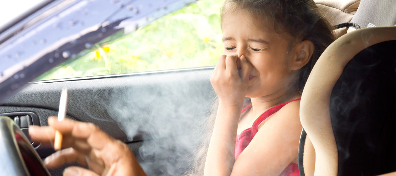 Ein Vater mit Zigarette sitzt mit seiner Tochter im Auto, die sich die Nase zuhält.
