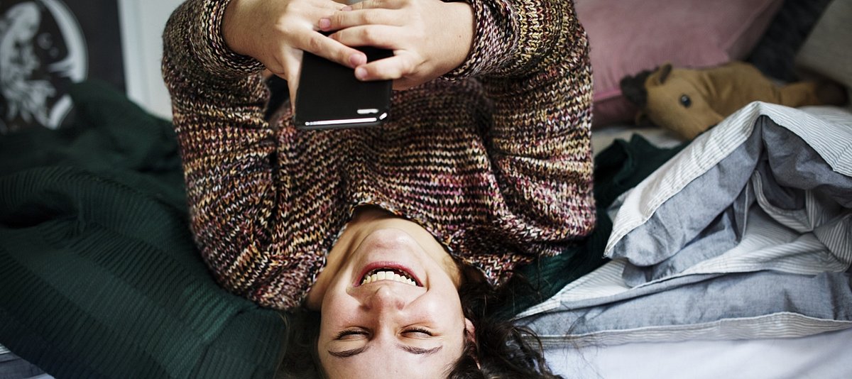 Ein Mädchen liegt auf ihrem Bett und hält lachend ihr Smartphone über sich und schreibt etwas