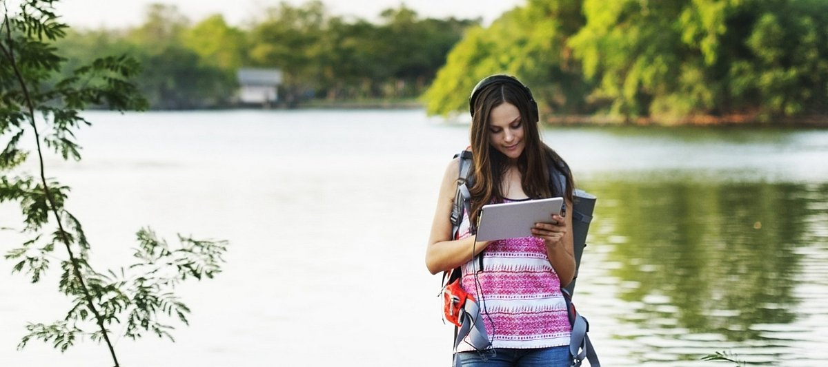 Eine Jugendliche mit Kopfhörer steht vor einem See und schaut auf ein Tablet