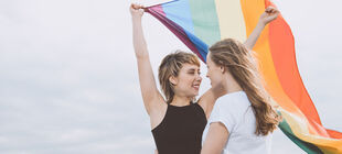 Zwei junge lesbische Frauen umarmen sich und eine hält eine Regenbogenfahne über sich.