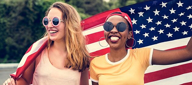 Zwei junge Frauen halten hinter ihrem Rücken eine USA-Flagge hoch und lachen im Sommer.