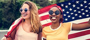 Zwei junge Frauen halten hinter ihrem Rücken eine USA-Flagge hoch und lachen im Sommer.