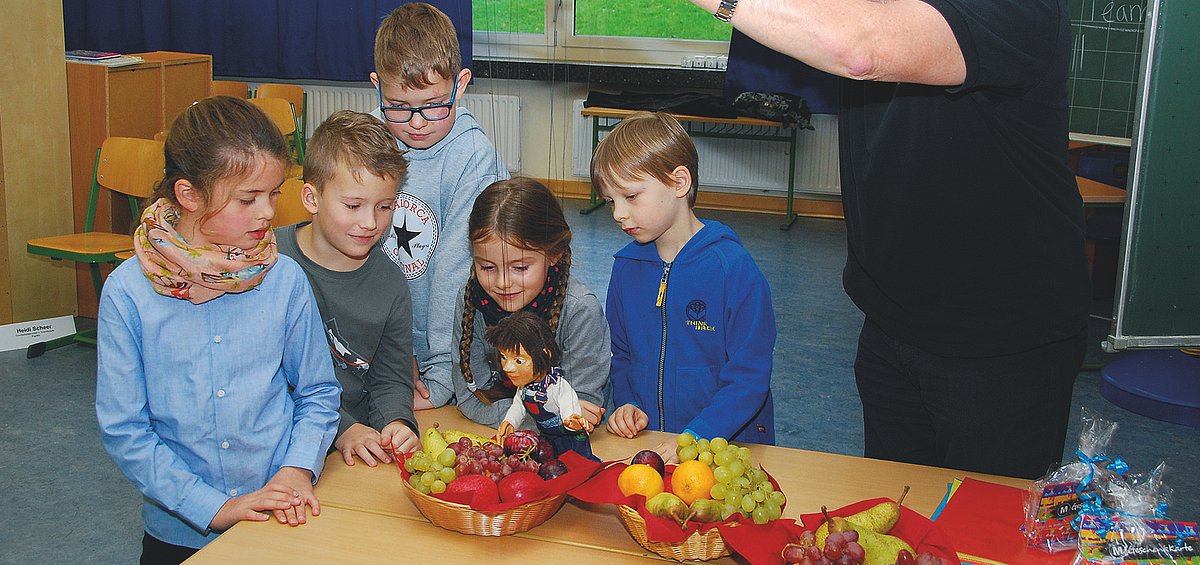 Grundschulkinder stehen um einen Tisch mit Obstkörben. Ein Mann hält eine Marionette über das Obst.