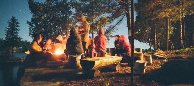 Jugendliche sitzen nach Sonnenuntergang auf Bänken im Kreis um ein Waldlagerfeuer.