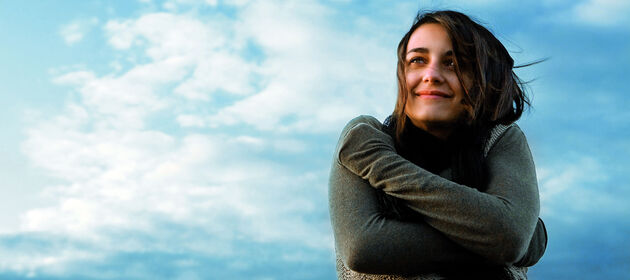 Lächelnde junge Frau umarmt sich selbst und steht vor einem blauen Himmel mit Wolken