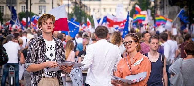 Eine junge Frau und ein junger Mann schauen in die Kamera, im Hintergrund eine Demonstration auf einer großen Stadtstraße. Menschen tragen Regenbogenflaggen, EU-Flaggen und polnische Flaggen.