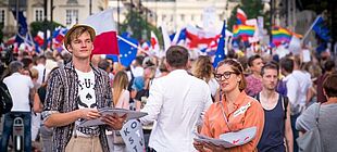 Eine junge Frau und ein junger Mann schauen in die Kamera, im Hintergrund eine Demonstration auf einer großen Stadtstraße. Menschen tragen Regenbogenflaggen, EU-Flaggen und polnische Flaggen.