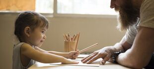 Ein Kind sitzt an einem Tisch und hat Stifte in der Hand, gegenüber sitzt ein Erwachsener