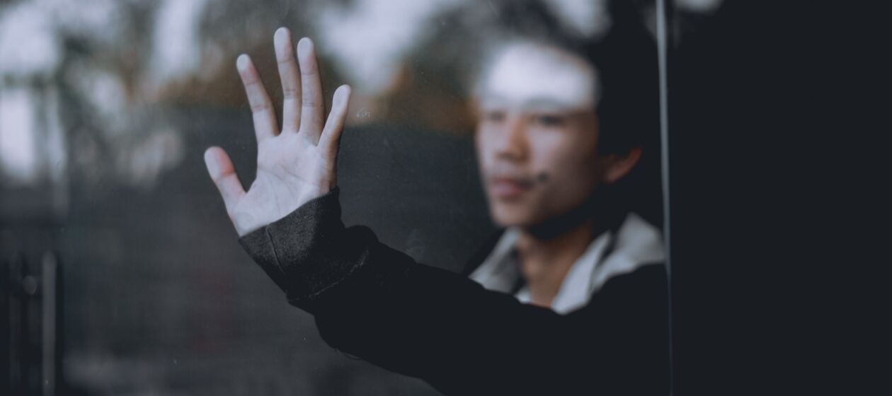 Ein Jugendlicher mit traurigem Gesichtsausdruck steht hinter einer Fensterscheibe und berührt mit ausgestreckter Hand das Glas