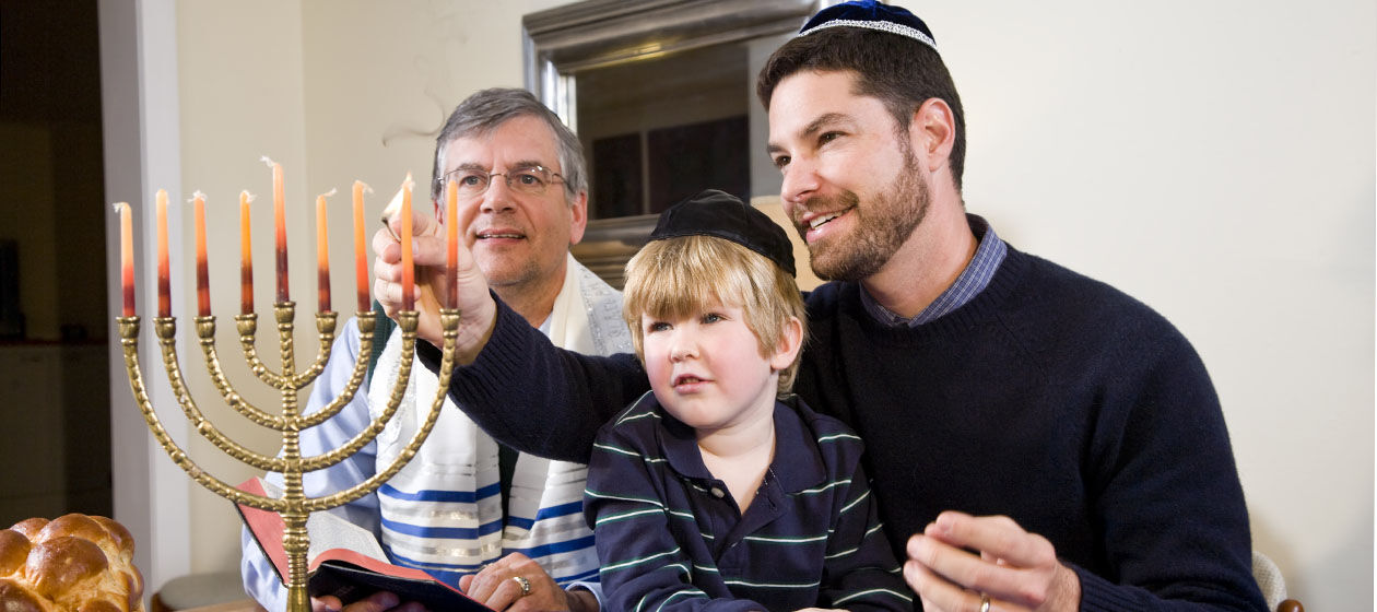 Ein jüdischer Vater zündet mit seinem Sohn und dem Großvater, der die Thora hält, die Kerzen am Chanukkaleuchter an.