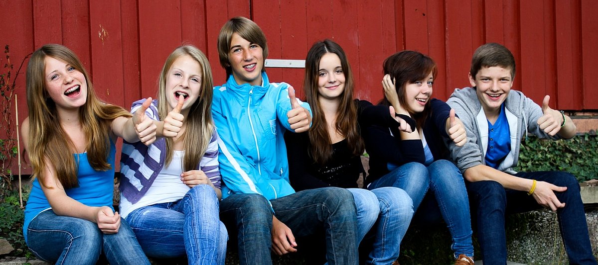Sechs lachende Jugendliche sitzen draußen vor einer Hütte mit herhobenen Daumen