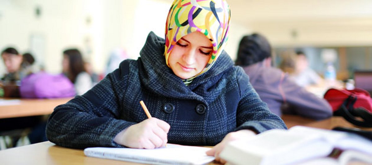 Eine junge Frau, die ein Kopftuch trägt, sitzt im Klassenzimmer und schreibt.