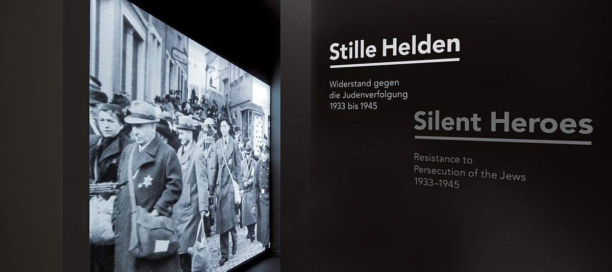 Eingang zur Ausstellung "Stille Helden" mit großformatigen Foto mit einer Straßenszene. Darauf zu sehen sind viele erwachsene Männer und Frauen, die der Judenstern auf ihren Mänteln und Jacken tragen. Im rechten Eck steht ein Polizeibeamter in Uniform. 