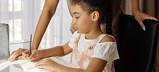 Ein Kind sitzt an einem Tisch und schreibt in ein Heft, eine erwachsene Person schaut ihm über die Schulter