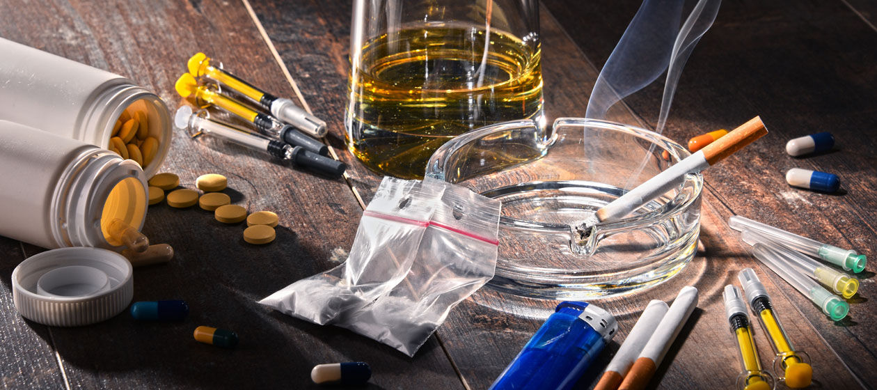 Neben einer brennenden Zigarette im Aschenbecher steht ein Glas mit einem alkoholischen Getränk, liegen zahlreiche Tabletten, Drogen und diverse Spritzen.