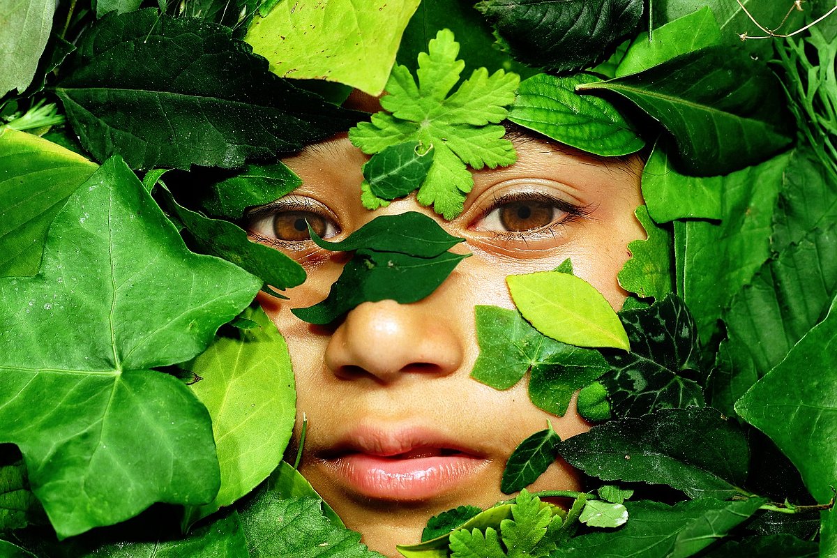 Die Fotografie zeigt ein junges Gesicht hinter vielen grünen Blättern.
