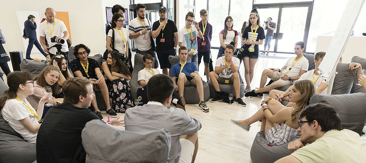 Jugendliche sitzen und stehen im Kreis in einem hellen Raum und diskutieren miteinander.