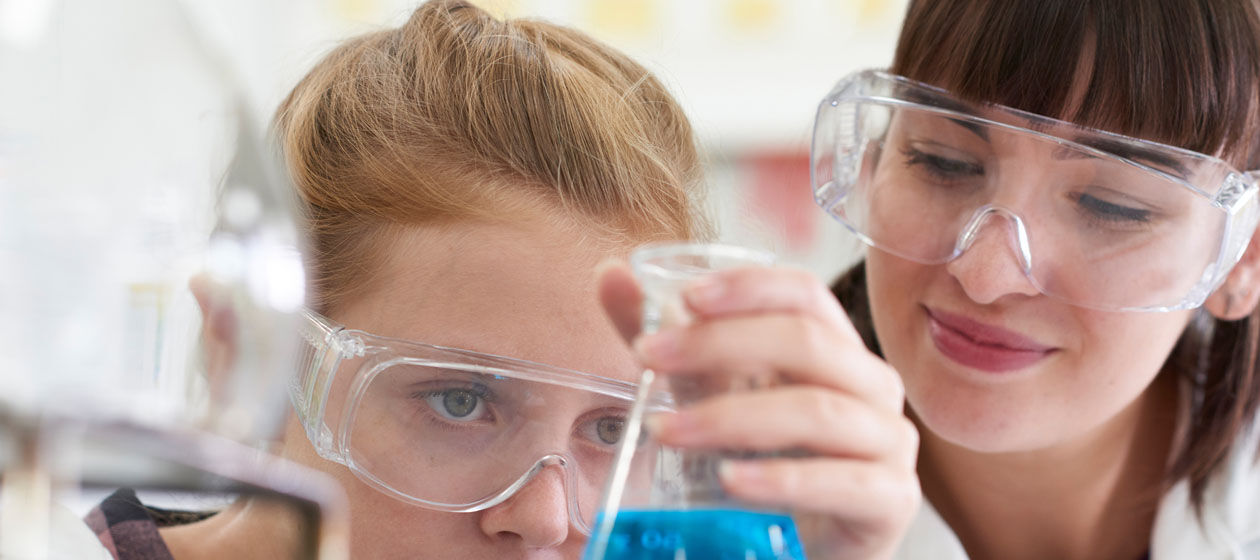 Eine Schülerin untersucht mit einer Lehrerin im Chemielabor eine blaue Flüssigkeit im Erlenmeyerkolben.