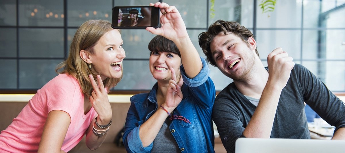 Drei junge Menschen sitzen vor einem Laptop und machen lachend ein Selfie