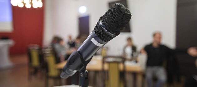 Eine Nahaufnahme eines Mikrofons in einem Mikrofonständer, im Hintergrund sind Personen an Tischen sitzend erkennbar