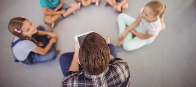 Mehrere Kinder sitzen in einem Kreis, eine erwachsene Person liest von einem Tablet vor