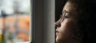 Junge Frau mit Brille schaut traurig aus dem Fenster.
