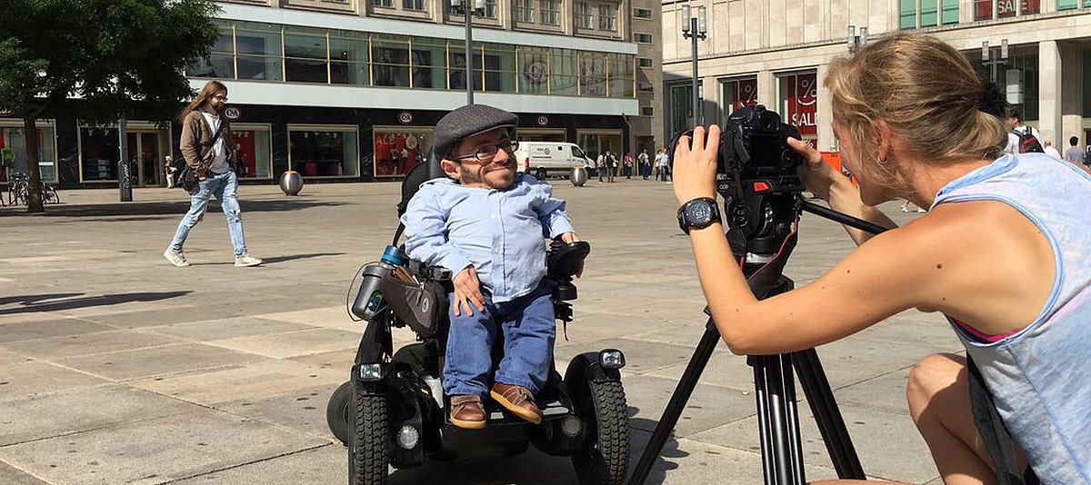 Raúl Krauthausen sitzt in seinem Rollstuhl auf dem Berliner Alexanderplatz und wird von einer Kamerafrau gefilmt. Im Hintergrund sieht man die Schaufenster der C&A-Filiale. 